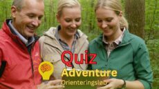 Billedet viser tre personer i en skov, der smilende kigger på en enhed, muligvis en tablet eller telefon, som en del af en aktivitet. Teksten "Quiz Adventure Orienteringsløb" er centralt placeret på billedet med en ikon af en lyspære og en hjerne, hvilket antyder en kombination af quiz og orienteringsløb. Deltagerne ser engagerede og glade ud, hvilket indikerer en sjov og interaktiv teambuilding-aktivitet i naturen.