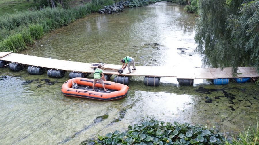 På billedet ser vi en teambuilding-aktivitet arrangeret af Bridge It. To deltagere bygger en flydende bro over en lavvandet flod ved hjælp af træplanker og tønder. Den ene deltager arbejder på broen, mens den anden holder en orange gummibåd.