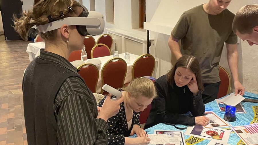 På billedet ser vi en gruppe mennesker, der deltager i en teambuilding-aktivitet med VR game (virtual reality) teknologi. En person bærer et VR-headset og holder en controller, mens de andre deltagere sidder ved et bord og arbejder sammen. På bordet ligger forskellige papirer, kort og dokumenter, som deltagerne ser ud til at analysere eller løse opgaver med. Aktiviteten foregår i et indendørs lokale