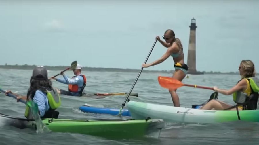 På billedet ser vi en gruppe mennesker, der deltager i kajaksejlads og stand-up paddleboarding på åbent vand. Tre personer sidder i kajakker, mens en person står op og padler. De bærer redningsveste og ser ud til at nyde aktiviteten