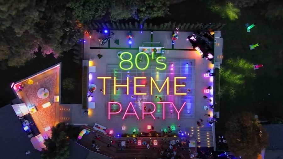 Billedet viser en farverig 80'er temafest set fra oven. Midt i billedet står teksten "80's THEME PARTY" skrevet i neonfarver. Festen foregår omkring en belyst tennisbane, omdannet til dansegulv, omgivet af lysende borde og siddeområder. Forskellige farvede lys skaber en festlig atmosfære, og træerne omkring området er også oplyst. Alt i alt viser billedet en energisk og festlig begivenhed i 80'er stil.