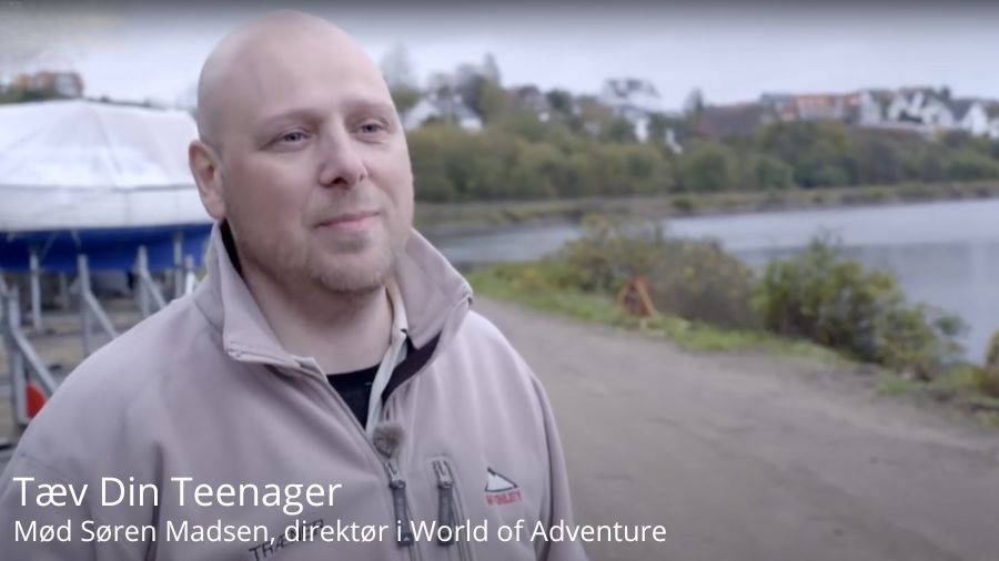 Billedet viser Søren Madsen, direktør i World of Adventure, stående ved en sø med både i baggrunden. I baggrunden ses en rolig søbred med træer og huse på den anden side af vandet. Teksten på billedet lyder "Tæv Din Teenager - Mød Søren Madsen, direktør i World of Adventure."