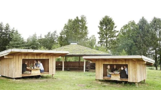 Billedet viser Haldbjerg Naturcampus, hvor man ser udendørs træhytter med borde og bænke samt en overdækket pavillon i baggrunden. Omgivet af frodige træer og grønne områder på Sjælland ser det ud til, at der er gode udendørsforhold til teambuilding-aktiviteter.