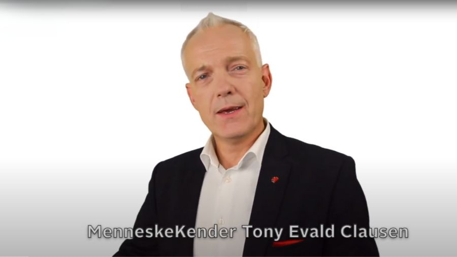 Billedet viser Tony Evald Clausen, der er kendt som "MenneskeKenderen". Han er iført en mørk jakke og en hvid skjorte, og han står foran en hvid baggrund. I øverste venstre hjørne af billedet er logoet og navnet "MenneskeKenderen" synligt. Teksten nederst på billedet lyder "MenneskeKender Tony Evald Clausen". Tony ser direkte ind i kameraet, hvilket giver et indtryk af, at han kommunikerer eller formidler en besked.