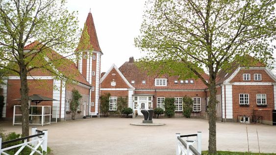 Billedet viser Sørup Herregaard, hvor man ser den røde murstensbygning med hvidmalede detaljer og et tårn. Bygningen er omgivet af en gårdsplads med træer og en indbydende atmosfære.