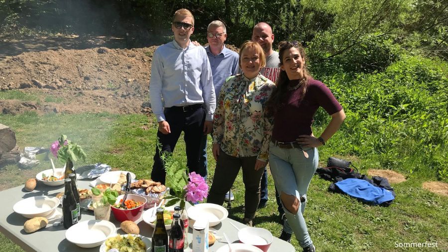 Billedet viser en gruppe mennesker, der deltager i en sommerfest. De står smilende omkring et bord fyldt med mad og drikkevarer i en grøn, udendørs omgivelser. Bordet er dækket med forskellige retter og drikkevarer, klar til at blive nydt. Teksten "Sommerfest" er diskret placeret nederst til højre på billedet.