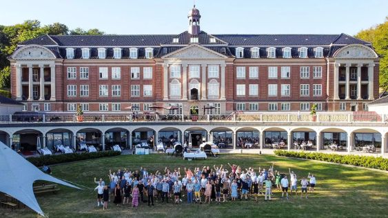 Billedet viser en stor gruppe mennesker samlet foran Hotel Koldingfjord, en imponerende bygning med klassisk arkitektur, beliggende ved Kolding Fjord. Deltagerne, som ser ud til at være en del af et teambuilding-event, står på en græsplæne foran hotellet og smiler og vinker til kameraet. Hotellets elegante facade med mange vinduer og en stor balkon danner en smuk baggrund for arrangementet. Teksten "Teambuilding-events" kan passende placeres i forbindelse med brugen af billedet.