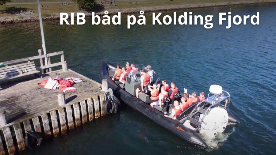 Billedet viser en RIB båd fyldt med deltagere i redningsveste, der sejler på Kolding Fjord. Båden er ved at lægge til ved en træbro, og deltagerne ser ud til at nyde oplevelsen. Teksten "Teambuilding-events RIB båd på Kolding Fjord" er centralt placeret på billedet.