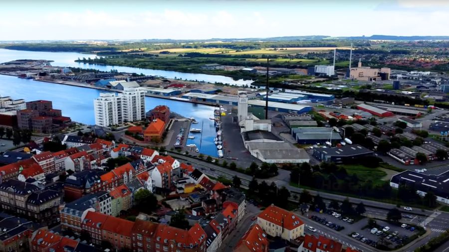 Billedet viser en udsigt over Horsens Havn og byen omkring den. I forgrunden ses byens tætte bebyggelse med røde tage, mens havnen med sine industrielle bygninger og skibe strækker sig ud mod vandet. I baggrunden kan man se grønne landskaber, der omgiver byen.