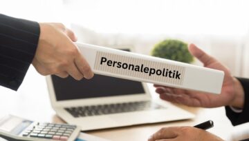 Billedet viser to personer i færd med at overdrage en mappe med etiketten "Personalepolitik". Baggrunden indeholder en laptop, en regnemaskine og en plante, hvilket indikerer et kontormiljø. Dette billede symboliserer vigtigheden af klare retningslinjer og politikker for medarbejdere på arbejdspladsen.