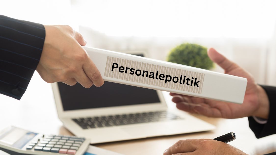 Billedet viser to personer i færd med at overdrage en mappe med etiketten "Personalepolitik". Baggrunden indeholder en laptop, en regnemaskine og en plante, hvilket indikerer et kontormiljø. Dette billede symboliserer vigtigheden af klare retningslinjer og politikker for medarbejdere på arbejdspladsen.